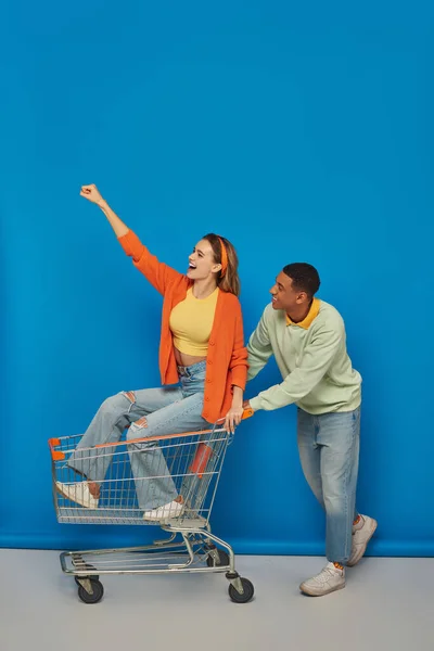 Positive african american man riding shopping cart with girlfriend inside of it on blue background — Stock Photo