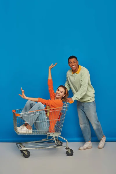 Freudig afrikanisch-amerikanischer Mann reitet Einkaufswagen mit glücklicher Freundin drinnen auf blauem Hintergrund — Stockfoto