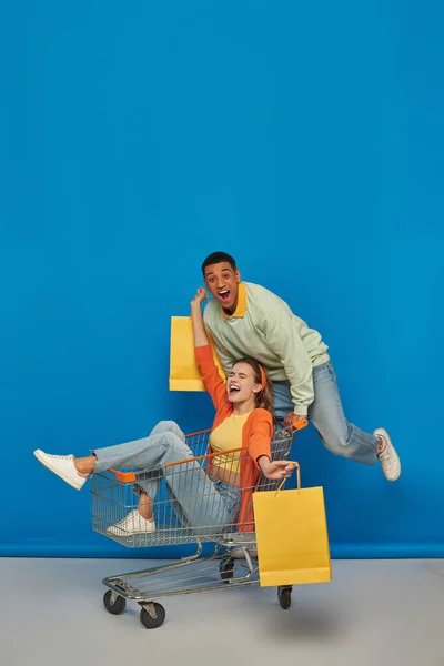 Glücklicher afrikanisch-amerikanischer Mann und freudige Frau im Einkaufswagen mit Einkaufstaschen auf blauem Hintergrund — Stockfoto