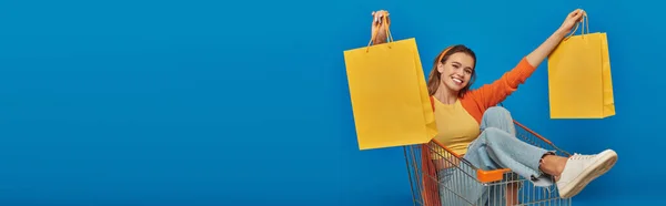 Happy woman sitting in cart and holding shopping bags on blue background, buying spree, banner — Stock Photo