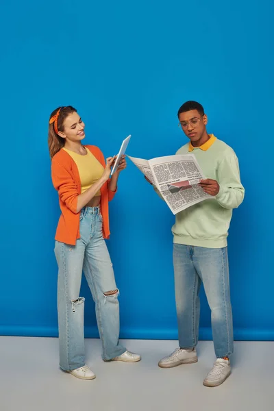 Mujer feliz usando la tableta cerca del hombre afroamericano en los anteojos que leen el periódico sobre fondo azul - foto de stock