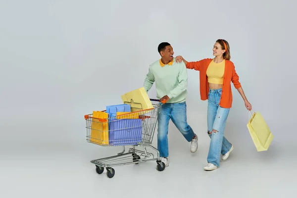 Animado interracial casal andando com carrinho e sacos de compras no fundo cinza, consumismo — Fotografia de Stock