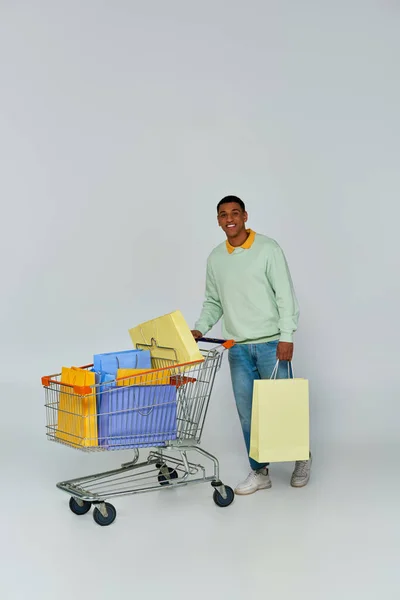 Animado afro-americano homem de pé com carrinho e sacos de compras em pano de fundo cinza, consumismo — Fotografia de Stock