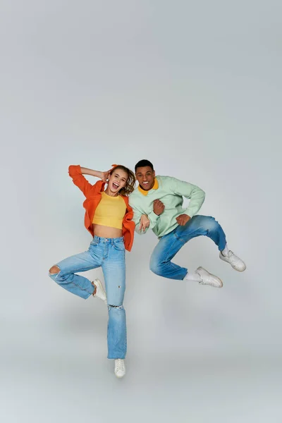 Excited interracial couple in casual attire jumping and looking at camera on grey backdrop, fun — Stock Photo