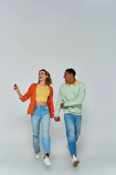 Glückliches interrassisches Paar in lässiger Kleidung, Händchen haltend und vor grauem Hintergrund zusammen laufend — Stockfoto