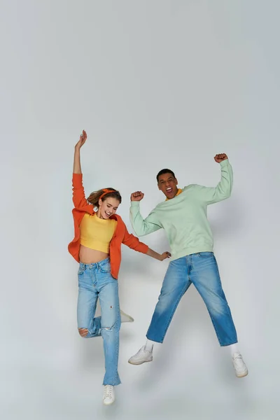 Feliz casal interracial em traje casual pulando juntos em pano de fundo cinza, espírito jovem — Fotografia de Stock