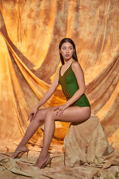 Modelo asiático bonito em saltos altos e maiô verde sentado na rocha, fundo bege com cortinas — Fotografia de Stock