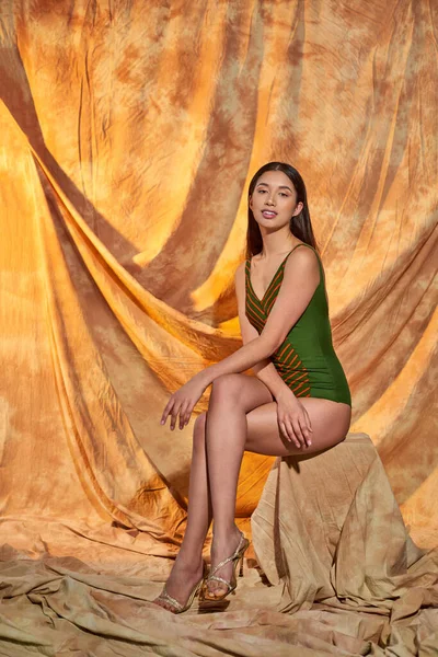 Cheerful asian model in high heels and green swimsuit sitting on beige background with drapery — Stock Photo