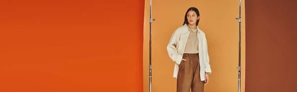 Bonita mujer asiática en traje de otoño posando con la mano en el bolsillo, pantalones de cuero y ropa de abrigo, bandera - foto de stock