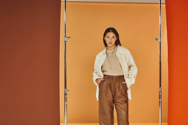 Mujer asiática joven en traje de otoño posando con las manos en los bolsillos, pantalones de cuero y ropa de abrigo - foto de stock