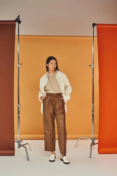 Morena mujer asiática en traje de otoño posando con las manos en los bolsillos, pantalones de cuero y ropa exterior - foto de stock