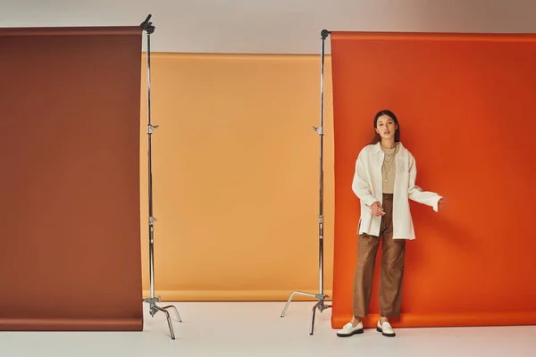 Стильная азиатская модель в осеннем наряде, стоящая на разноцветном фоне, кожаных брюках и верхней одежде — стоковое фото