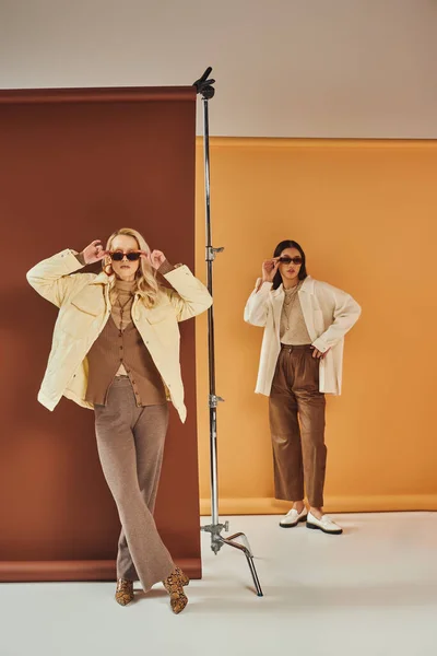 Fall season, interracial women in sunglasses and autumn outerwear posing on duo color backdrop — Stock Photo