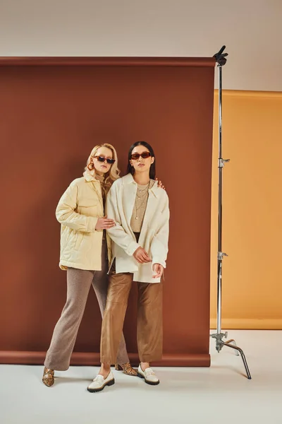 Temporada de outono, mulheres inter-raciais em óculos de sol e outerwear posando juntos no cenário de cor duo — Fotografia de Stock