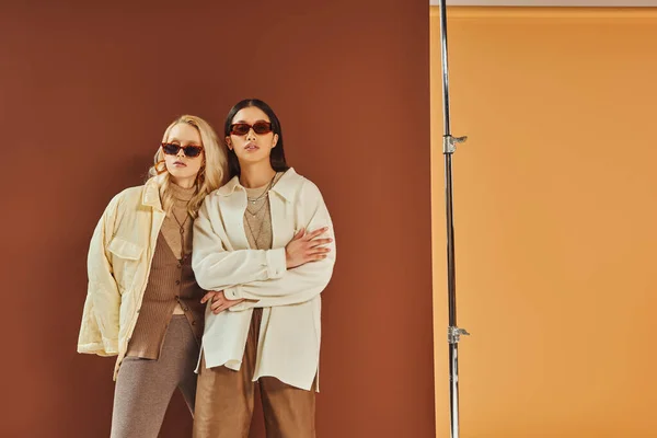 Осенняя мода и тенденции, межрасовые женщины в солнцезащитных очках и верхней одежде позируют на фоне цвета дуэта — стоковое фото