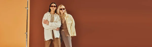 Fall trends, interracial women in sunglasses and outerwear posing on duo color backdrop, banner — Stock Photo