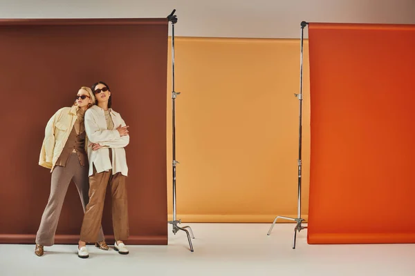 Temporada otoñal, modelos multiétnicos en gafas de sol y ropa de abrigo de otoño posando sobre un colorido telón de fondo - foto de stock