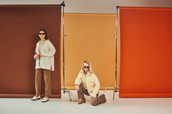 Interracial models in sunglasses and autumn outerwear posing on colorful backdrop, fall colors — Stock Photo