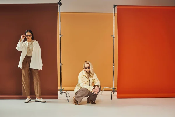 Donne interrazziale in occhiali da sole e capispalla autunno in posa su sfondo colorato, moda autunno — Foto stock