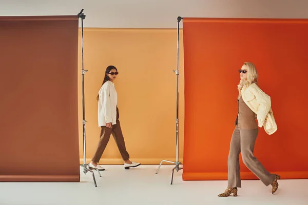 Разнообразные культуры женщины в верхней одежде и солнцезащитных очках ходить рядом красочный фон, осень моды — стоковое фото