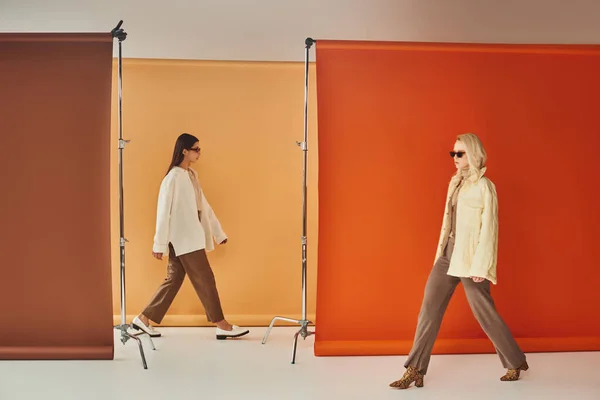Multiculturais mulheres em Outono outerwear e óculos de sol andando perto de pano de fundo colorido, moda queda — Fotografia de Stock