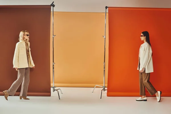 Modelos inter-raciais em outerwear e óculos de sol andando perto de pano de fundo colorido, conceito de moda queda — Fotografia de Stock