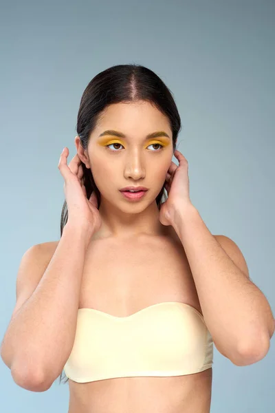 Asiatisches Model mit fettem Make-up posiert in trägerlosem BH vor blauem Hintergrund, strahlende Haut — Stockfoto