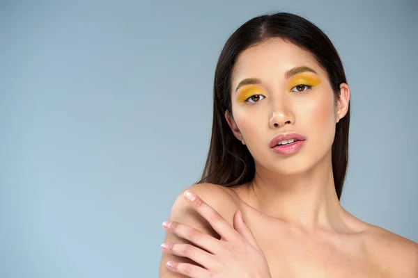 Selbstdarstellung, junges asiatisches Model mit kühnem Make-up und nackten Schultern posiert vor blauem Hintergrund — Stockfoto