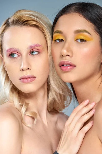 Concepto de belleza diversa, mujeres multiétnicas con maquillaje de ojos vibrante mirando a la cámara en el fondo azul - foto de stock