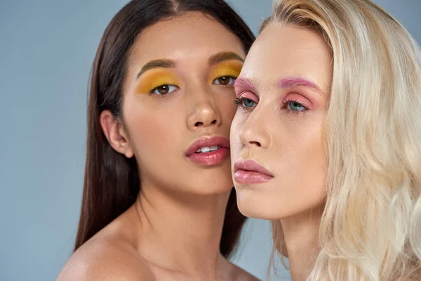 Interracial women with vibrant eye makeup posing together on blue backdrop, expressive eyes — Stock Photo