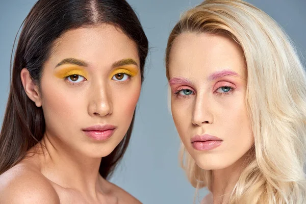 Modelos interraciales con maquillaje de ojos en negrita posando juntos sobre fondo azul, concepto de tendencia de belleza - foto de stock