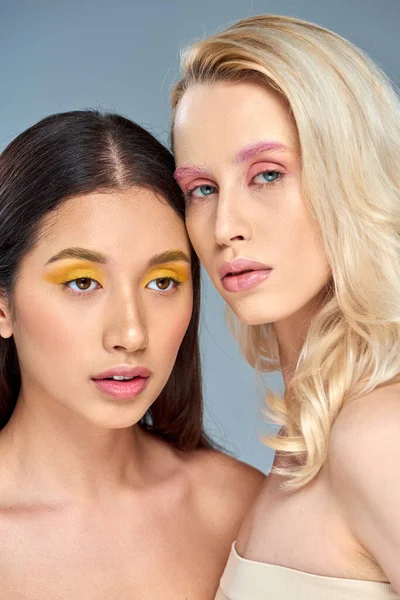 Interracial jóvenes mujeres con negrita ojo maquillaje posando juntos en azul telón de fondo, belleza tendencia concepto - foto de stock