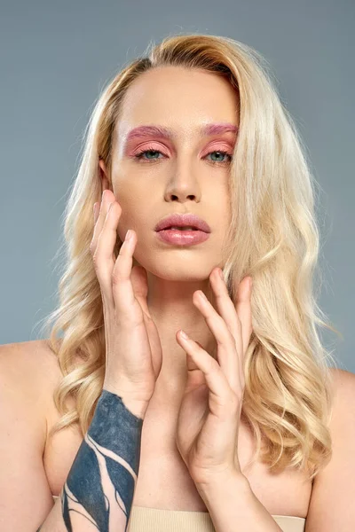 Tätowierte Frau mit fettem Augen-Make-up und blonden Haaren posiert vor grauem Hintergrund, feminine Schönheit — Stockfoto