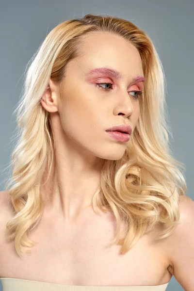 Femme sensuelle avec maquillage des yeux rose et cheveux blonds regardant loin sur fond gris, beauté féminine — Photo de stock