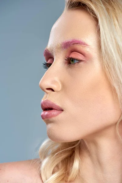 Primer plano de modelo con maquillaje de ojos rosados y cabello rubio posando sobre fondo gris, belleza femenina - foto de stock