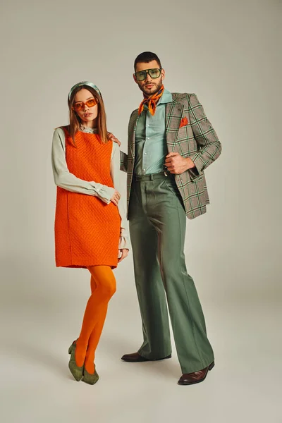 Jeune couple en tenue vintage élégante et lunettes de soleil debout sur fond gris, pleine longueur — Photo de stock