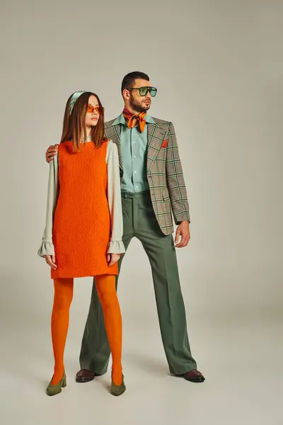 Full length of man in plaid blazer and woman in orange dress looking away on grey, retro fashion — Stock Photo