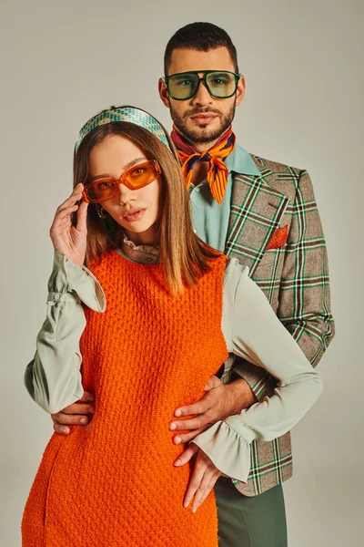 Mann im karierten Blazer umarmt Frau in orangefarbenem Kleid und Sonnenbrille auf grauer, retro-inspirierter Mode — Stockfoto