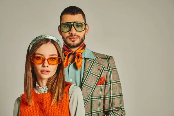 Молода пара в елегантному вінтажному одязі і сонцезахисних окулярах дивиться на камеру на сірому, мода з минулого — Stock Photo