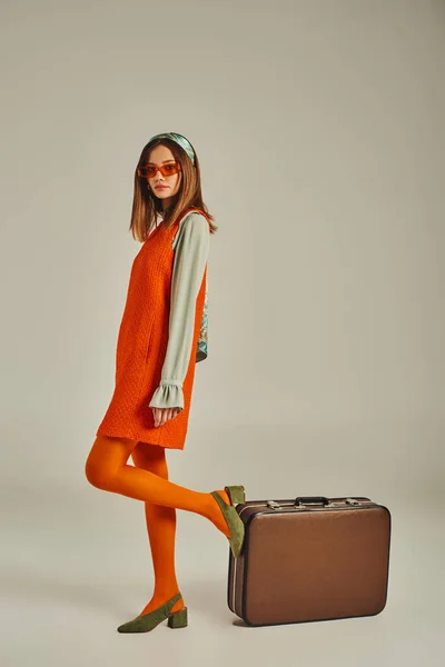 Trendy woman in orange dress and sunglasses posing near vintage suitcase on grey, retro style — Stock Photo