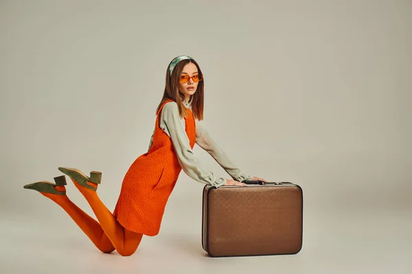 Young woman in stylish retro clothes and sunglasses kneeling near vintage suitcase on grey — Stock Photo