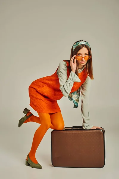 Гламурная женщина в ретро-одежде регулирует модные солнцезащитные очки и позирует рядом с винтажным чемоданом на сером — стоковое фото