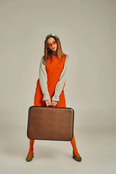 Femme de style rétro en robe orange et lunettes de soleil tendance avec valise vintage sur gris, pleine longueur — Photo de stock
