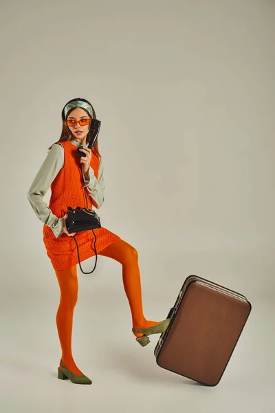 Молодая старомодная женщина в солнечных очках разговаривает по ретро-ротационному телефону возле винтажного чемодана на сером — стоковое фото