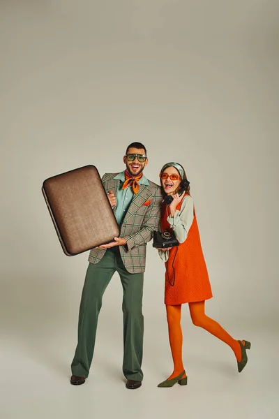 Весёлый мужчина с винтажным чемоданом рядом с женщиной разговаривает по ретро-телефону на серой, старомодной паре — стоковое фото