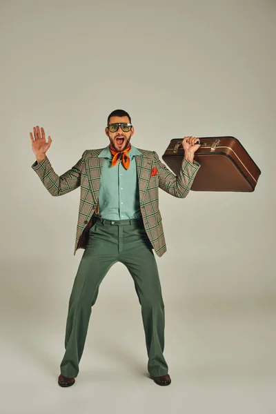 Fröhlicher Mann in karierter Jacke und Sonnenbrille mit Vintage-Koffer über grauen, retro-inspirierten Lebensstil — Stockfoto