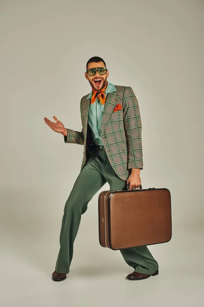 Uomo allegro in giacca a scacchi e occhiali da sole in posa con valigia vintage su grigio, stile retrò — Foto stock