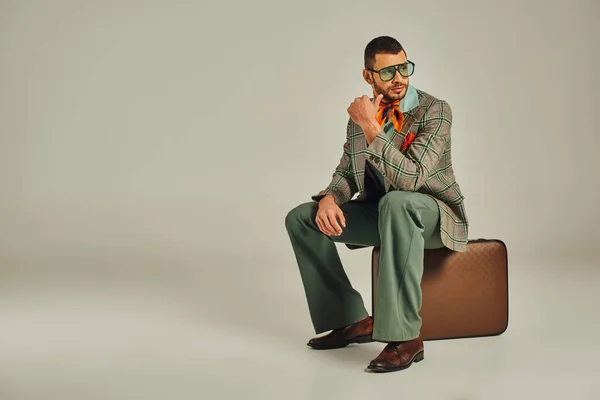 Uomo riflessivo stile vintage in occhiali da sole seduto sulla valigia retrò e guardando altrove sul grigio — Foto stock
