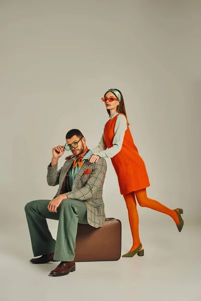 Frau in orangefarbenem Kleid neben stilvollem Mann auf Vintage-Koffer auf grauen, altmodischen Reisenden sitzend — Stockfoto
