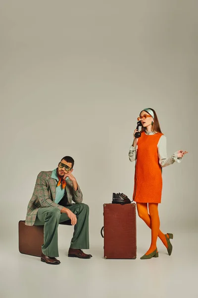 Женщина в оранжевом платье разговаривает по беспроводному телефону рядом со скучающим мужчиной, сидящим на винтажном чемодане на сером — стоковое фото
